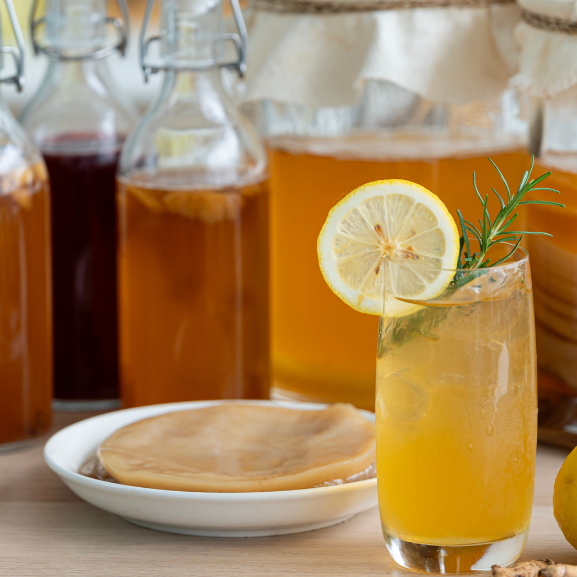Healthy kombucha SCOBY in a bowl, next to a glass jar of kombucha with lemon and rosemary, surrounded by large bottles of fermenting kombucha.