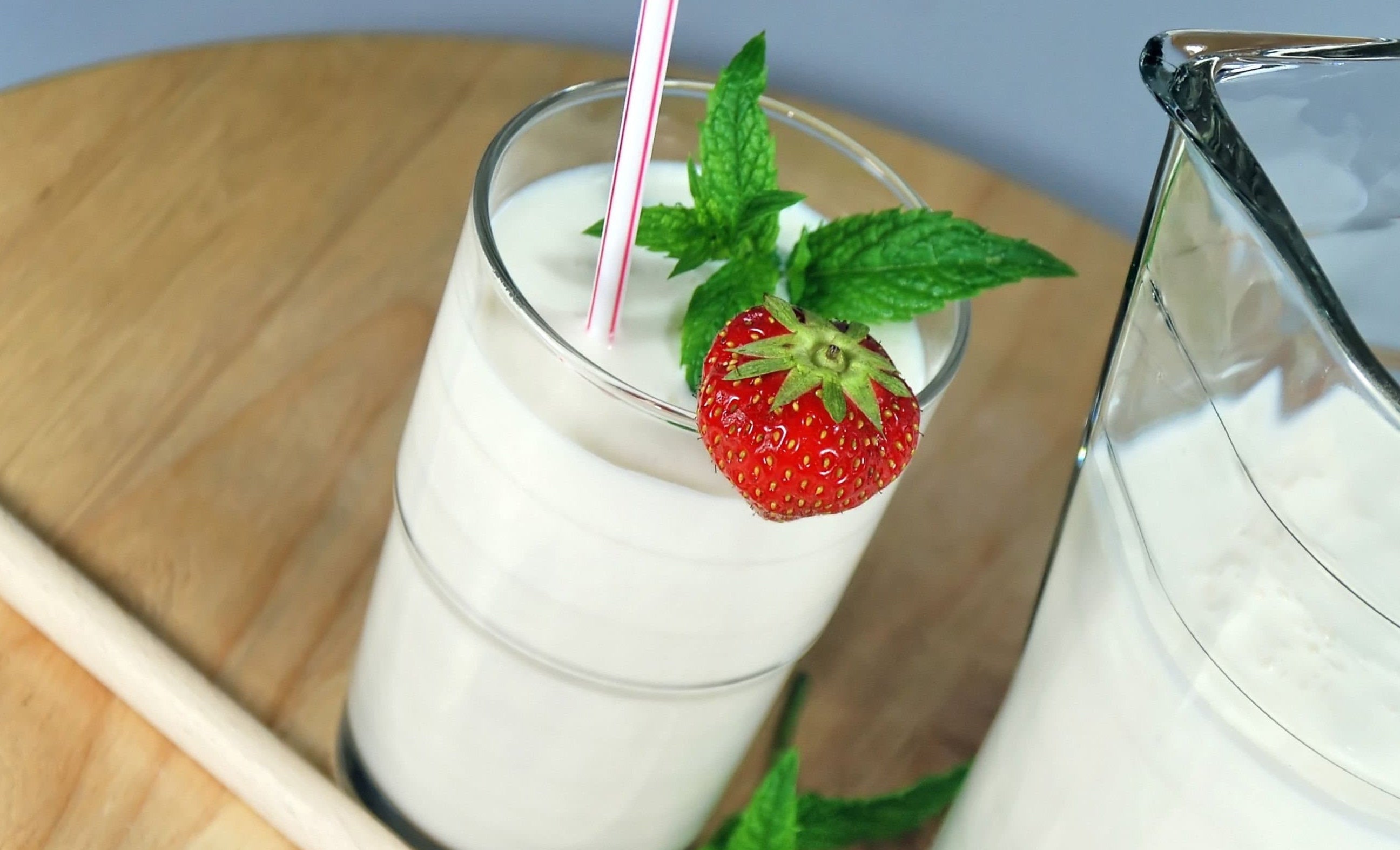 Glass of milk kefir with a strawberry and mint leaf, next to a wooden spoon with kefir grains and additional strawberries on a wooden surface.