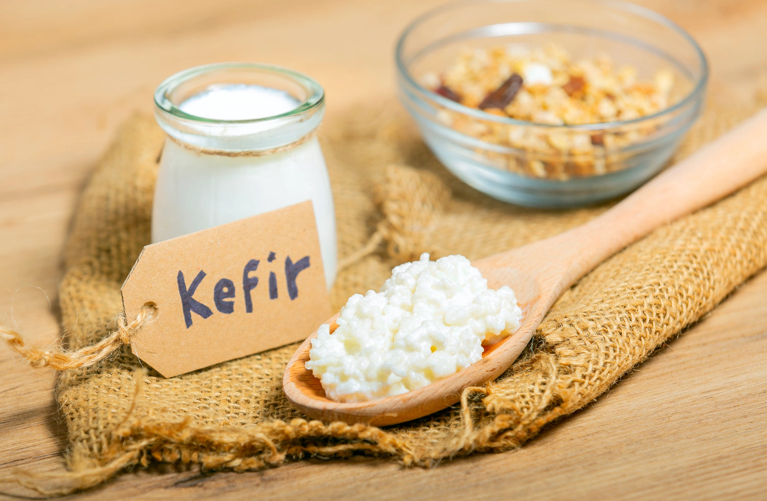 Fresh milk kefir grains on a wooden spoon, placed beside a bowl of nuts and a bottle of homemade probiotic milk kefir.