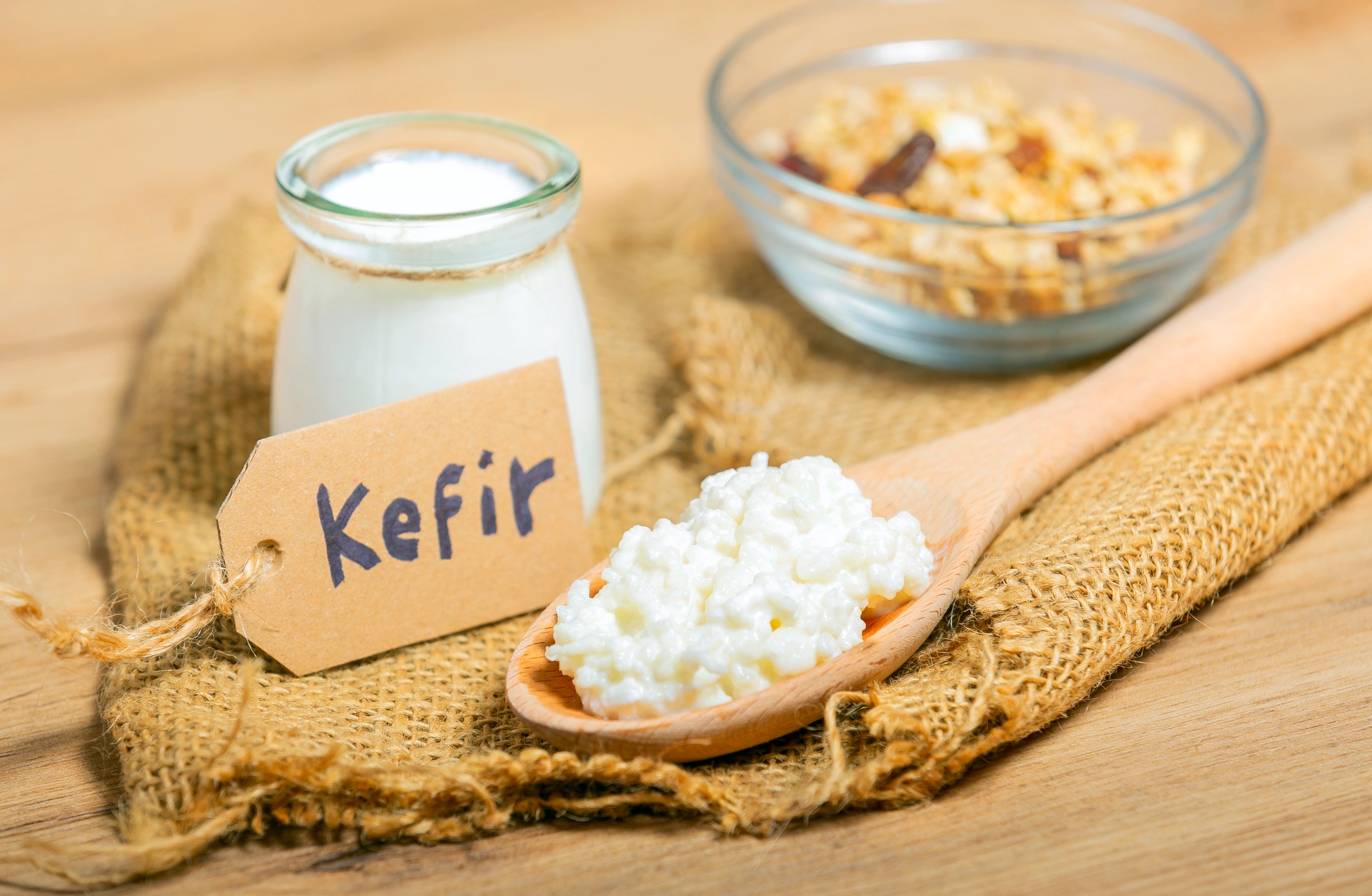 Fresh milk kefir grains on a wooden spoon, placed beside a bowl of nuts and a bottle of homemade probiotic milk kefir.
