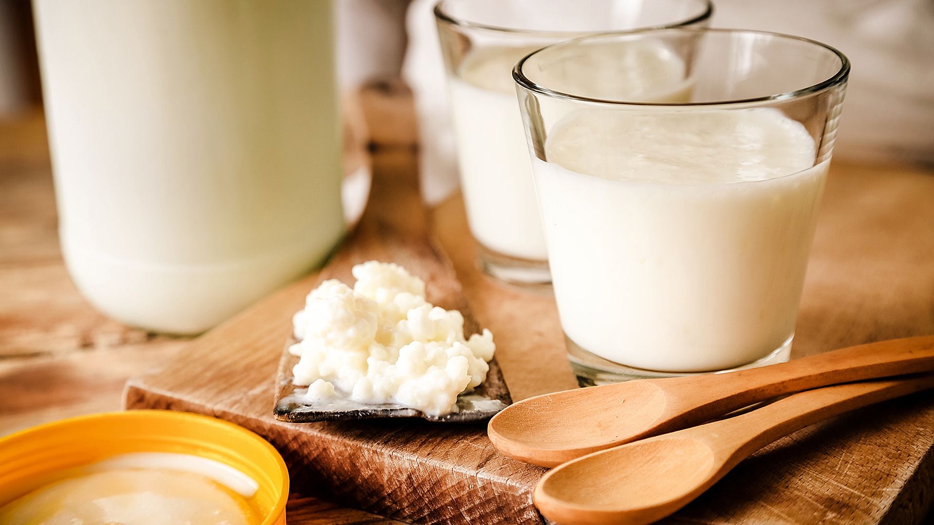 Two glasses of milk kefir with large jug of milk kefir in background sitting on top of wooden cutting board next to two wooden spoons and a pile of milk kefir grains.