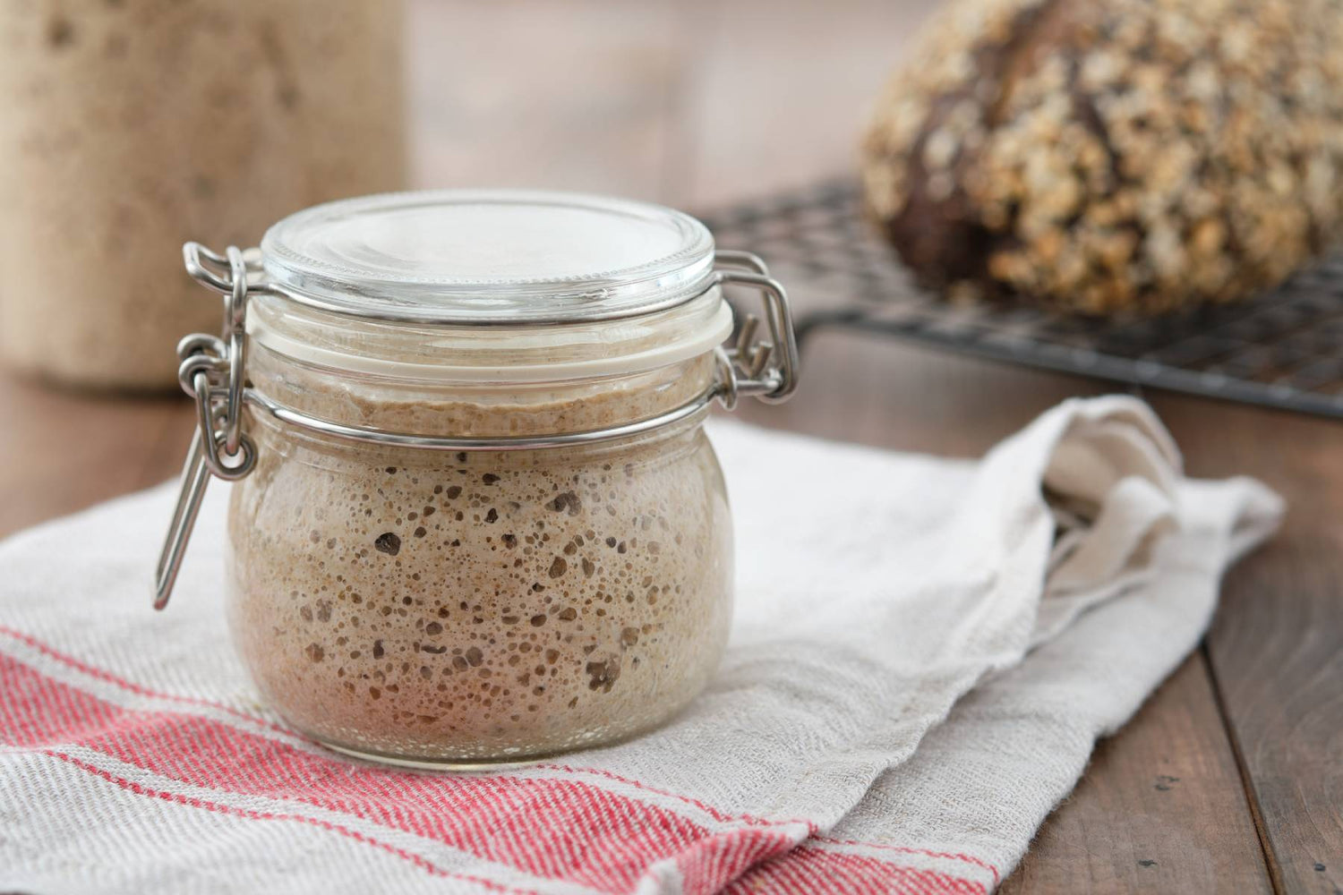 Bubbling sourdough starter in a jar, resting on a cloth with freshly baked bread in the background.