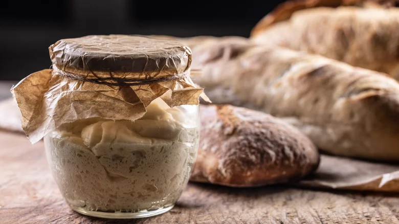 Homemade sourdough starter in a glass jar with rustic artisan bread loaves in the background on a wooden table.