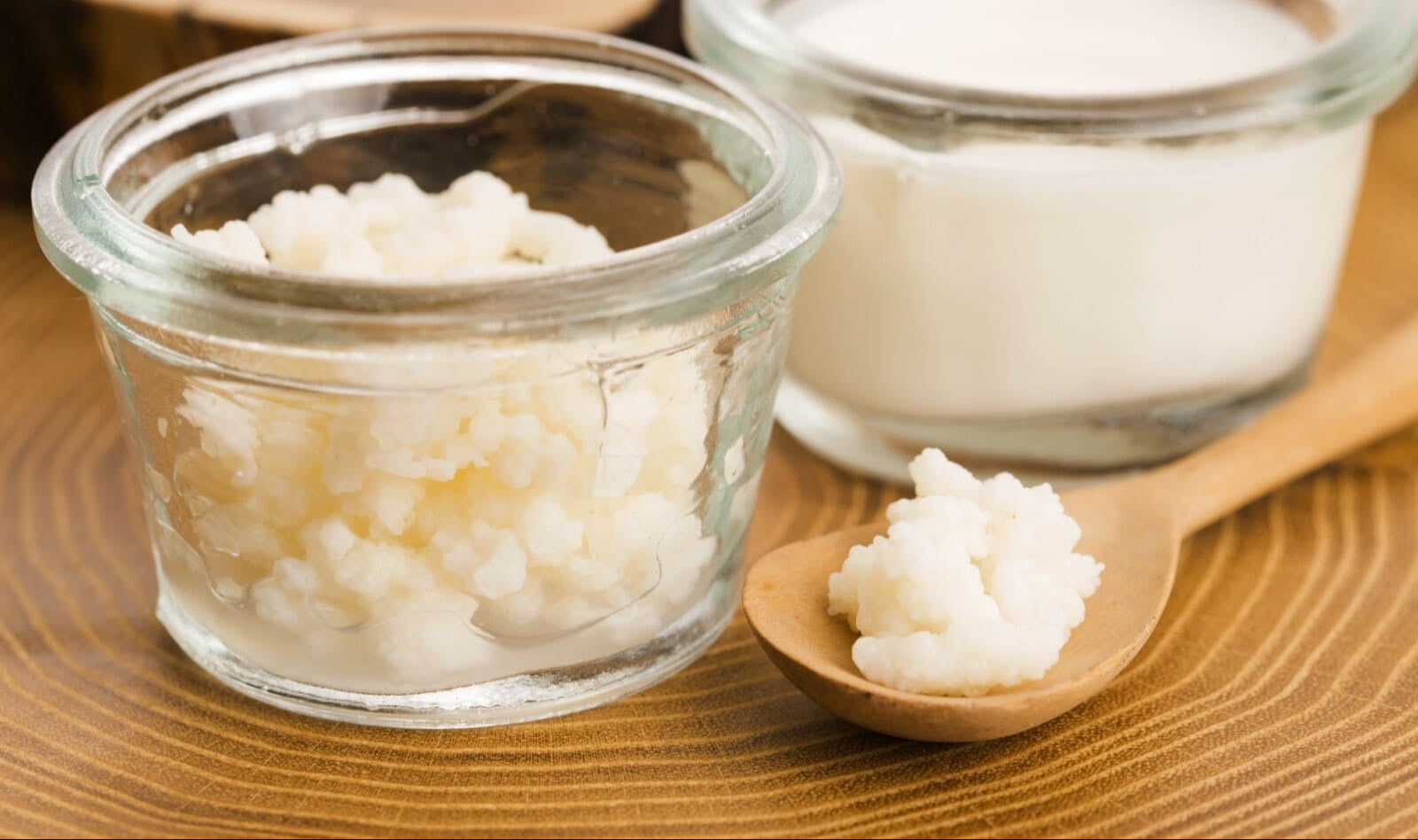 Two glass jars -- one with milk kefir, one with milk kefir grains, sitting on rustic surface next to wooden spoon holding milk kefir grains.