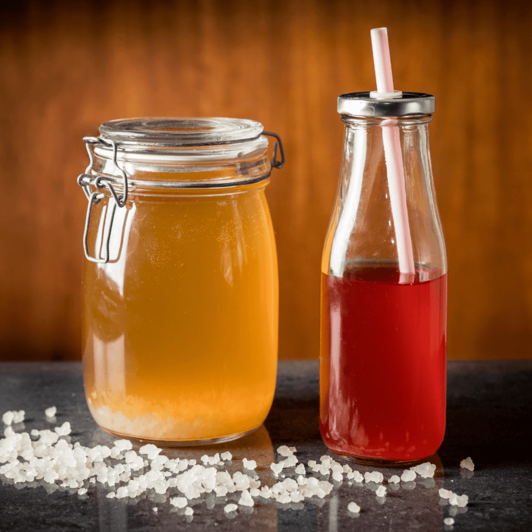 A jar of fermenting water kefir next to a glass jar of flavoured water kefir on table with kefir grains spread out in front.