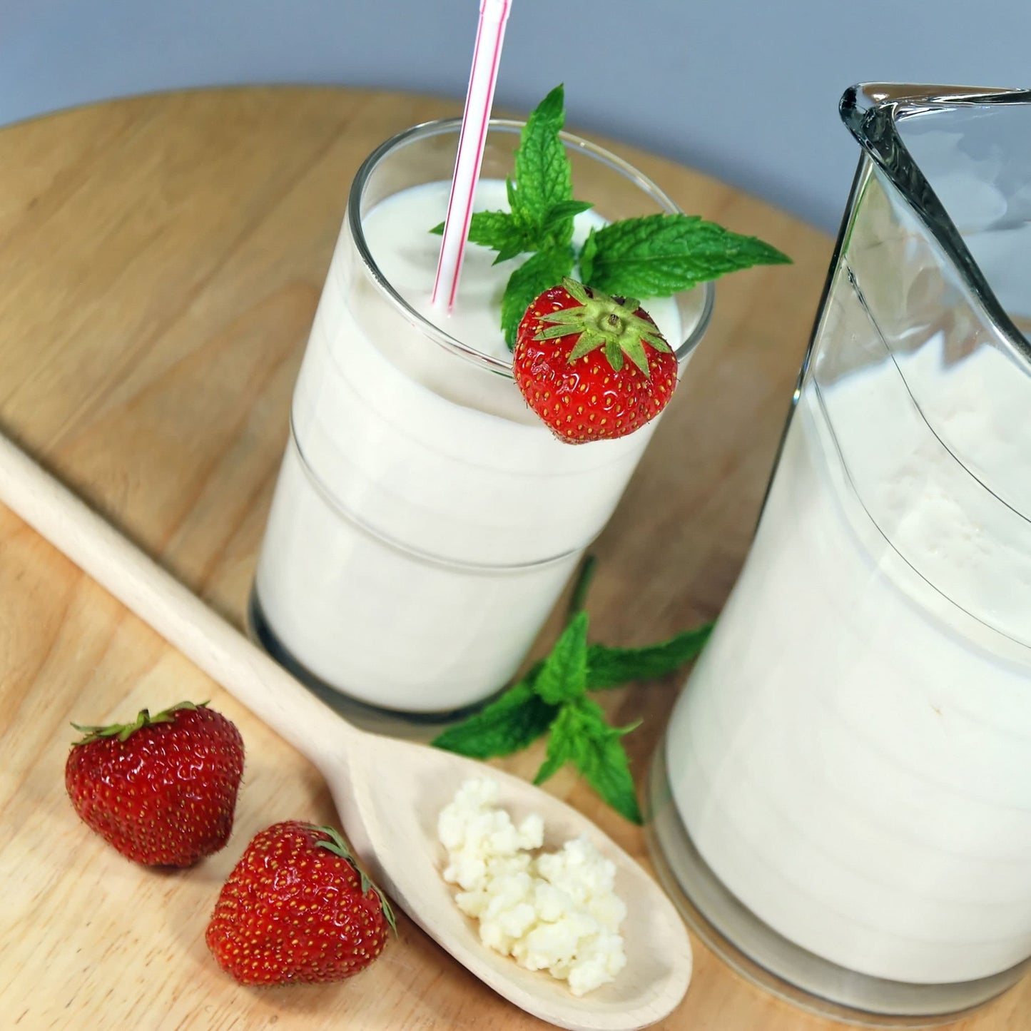 Fresh homemade milk kefir served in a glass with strawberries and mint, next to a wooden spoon with kefir grains and a glass jug filled with probiotic kefir drink.