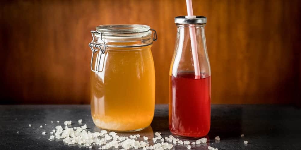Glass Jar of Fermenting Water kefir Next to Bottle of Water Kefir With Straw  Sitting on Table with Pile of Water Kefir Grains